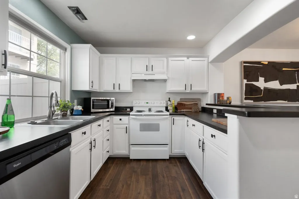 U-shaped kitchen featuring white cabinetry, dark countertops, wood-finish flooring, a stainless steel dishwasher, and a window above the sink