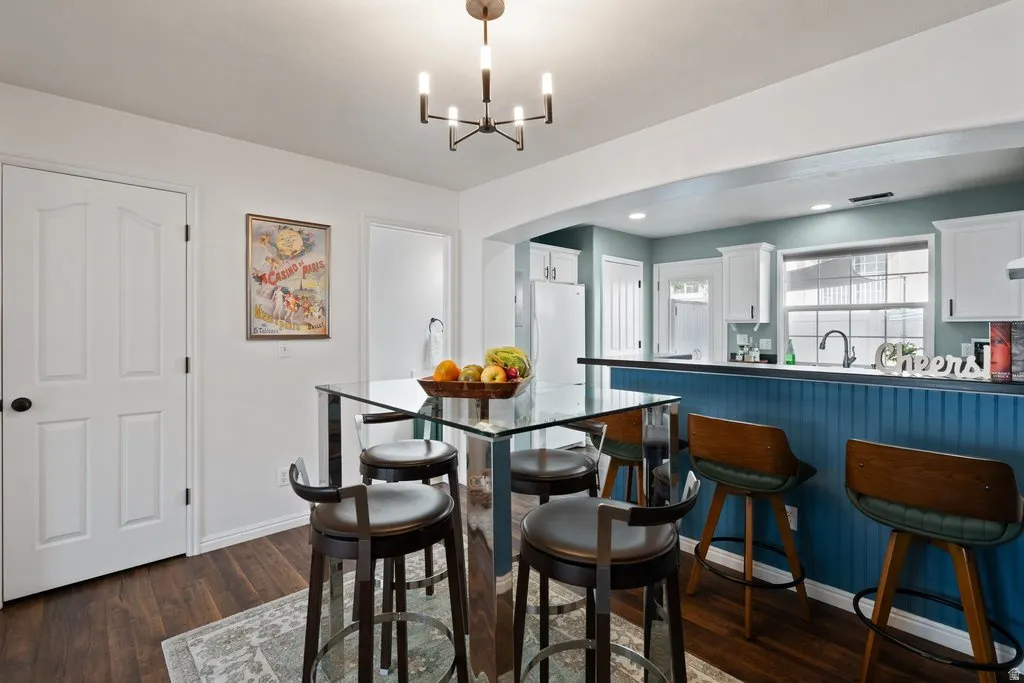 Modern kitchen area featuring a central glass-top table, wood-finish flooring, a contemporary chandelier, white cabinetry, and a blue paneled breakfast bar