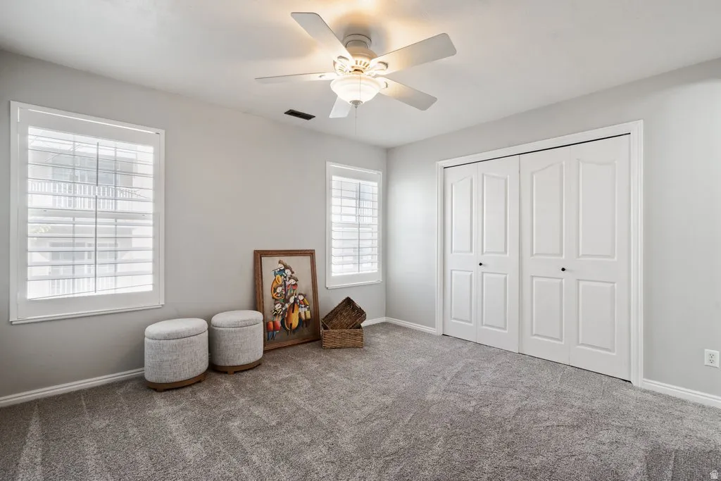 Carpeted interior room featuring two windows with white plantation shutters, a ceiling fan with integrated lighting, and a bi-fold closet with paneled doors