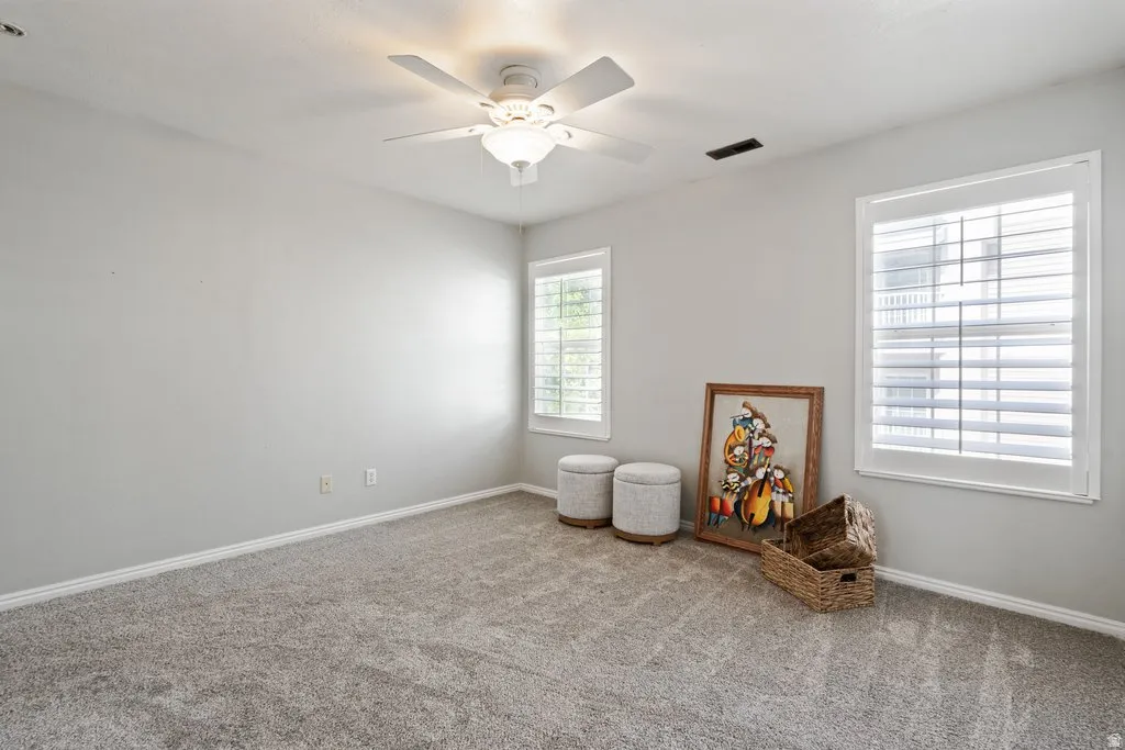 Bright interior space featuring two windows with white plantation shutters, a ceiling fan with integrated lighting, and neutral-toned carpeting