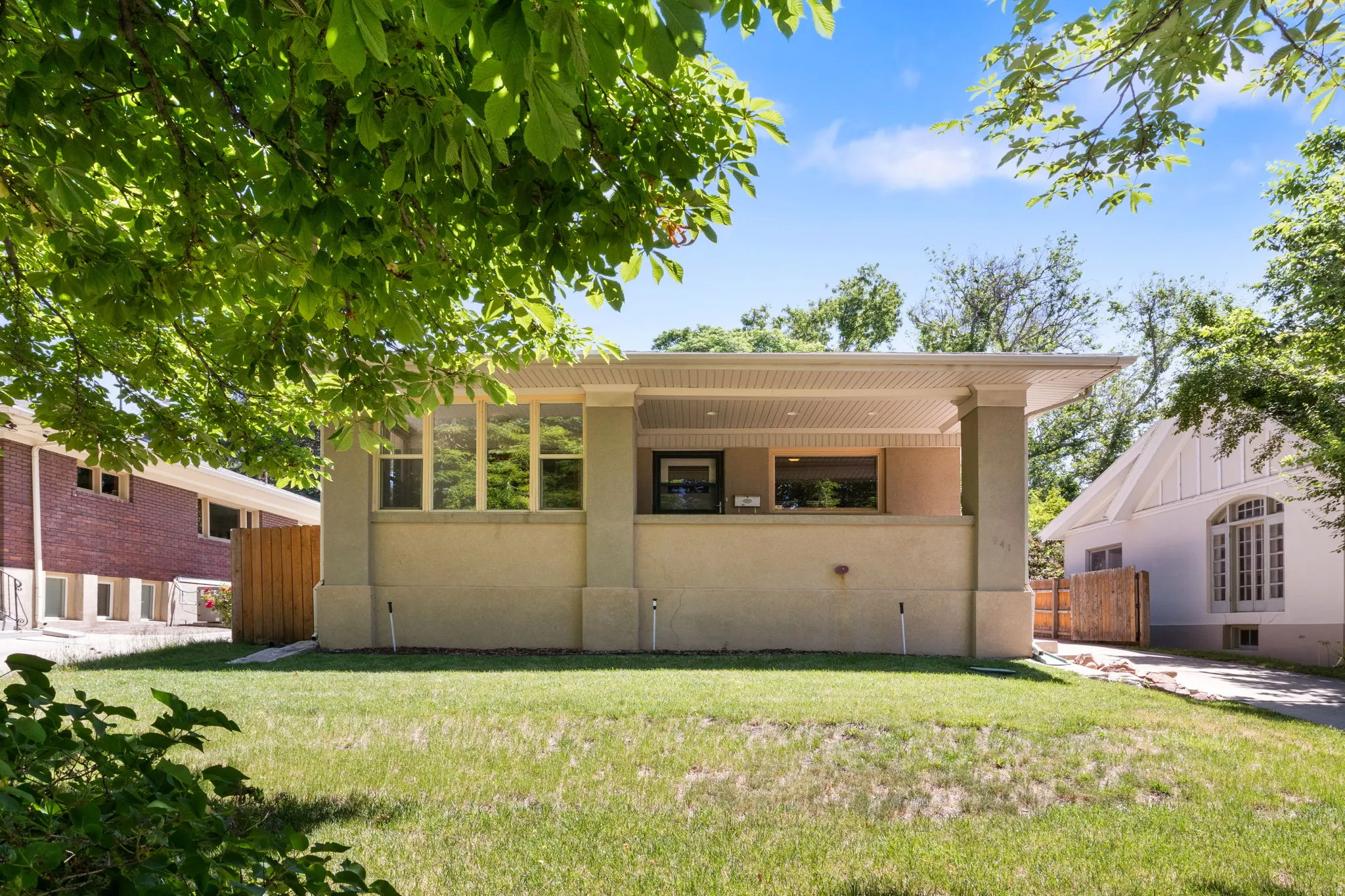 Single-story stucco exterior featuring a covered front porch with substantial columns, a front lawn, and a light wood privacy fence
