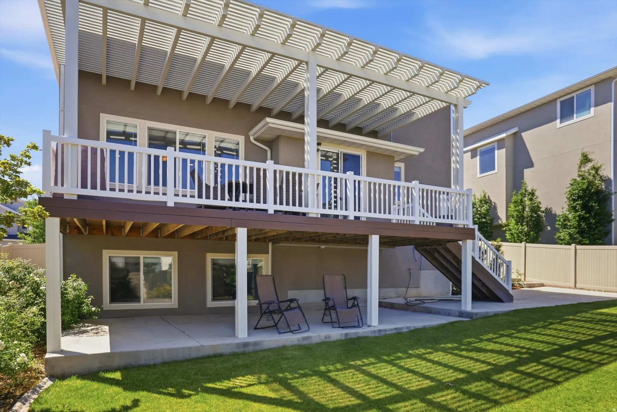 Two-story exterior featuring a private upper deck with white railing and a louvered pergola, a lower patio, and a manicured lawn