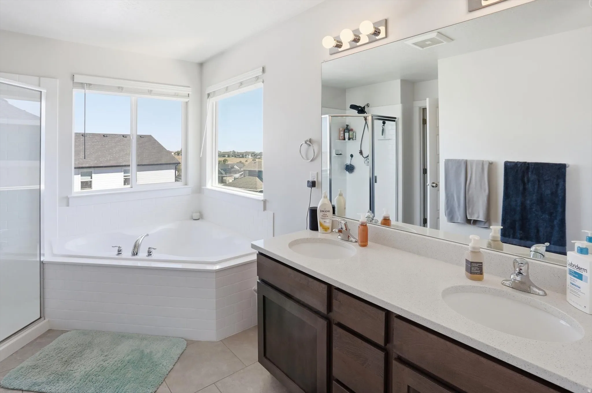 Bathroom featuring a corner soaking tub, separate glass-enclosed shower, dual vanity with undermount sinks, wood-finish cabinetry, and a large mirror with integrated lighting