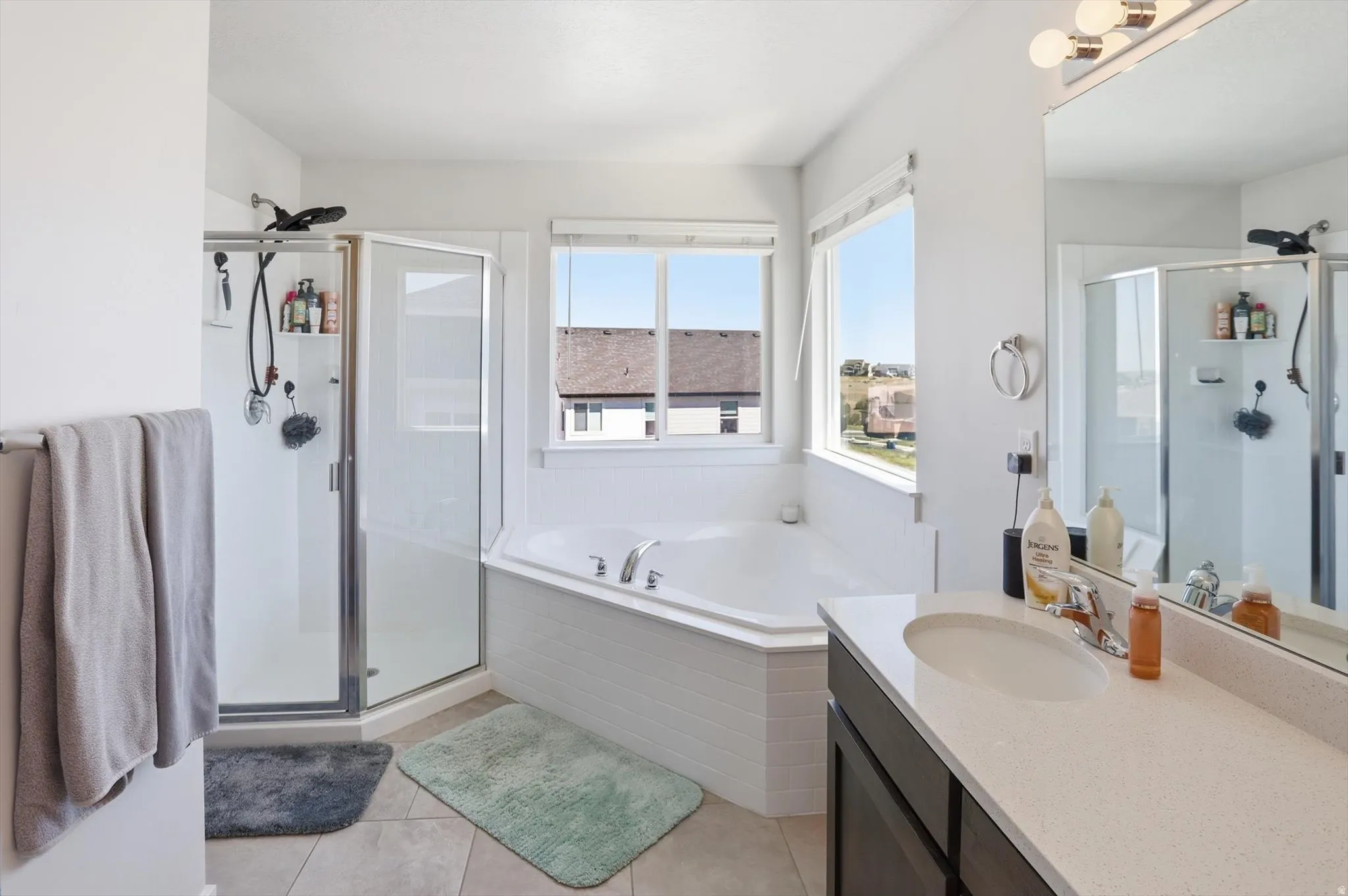 Bathroom featuring a built-in corner soaking tub, separate glass-enclosed shower, single vanity with a solid surface countertop, and large mirror with overhead lighting