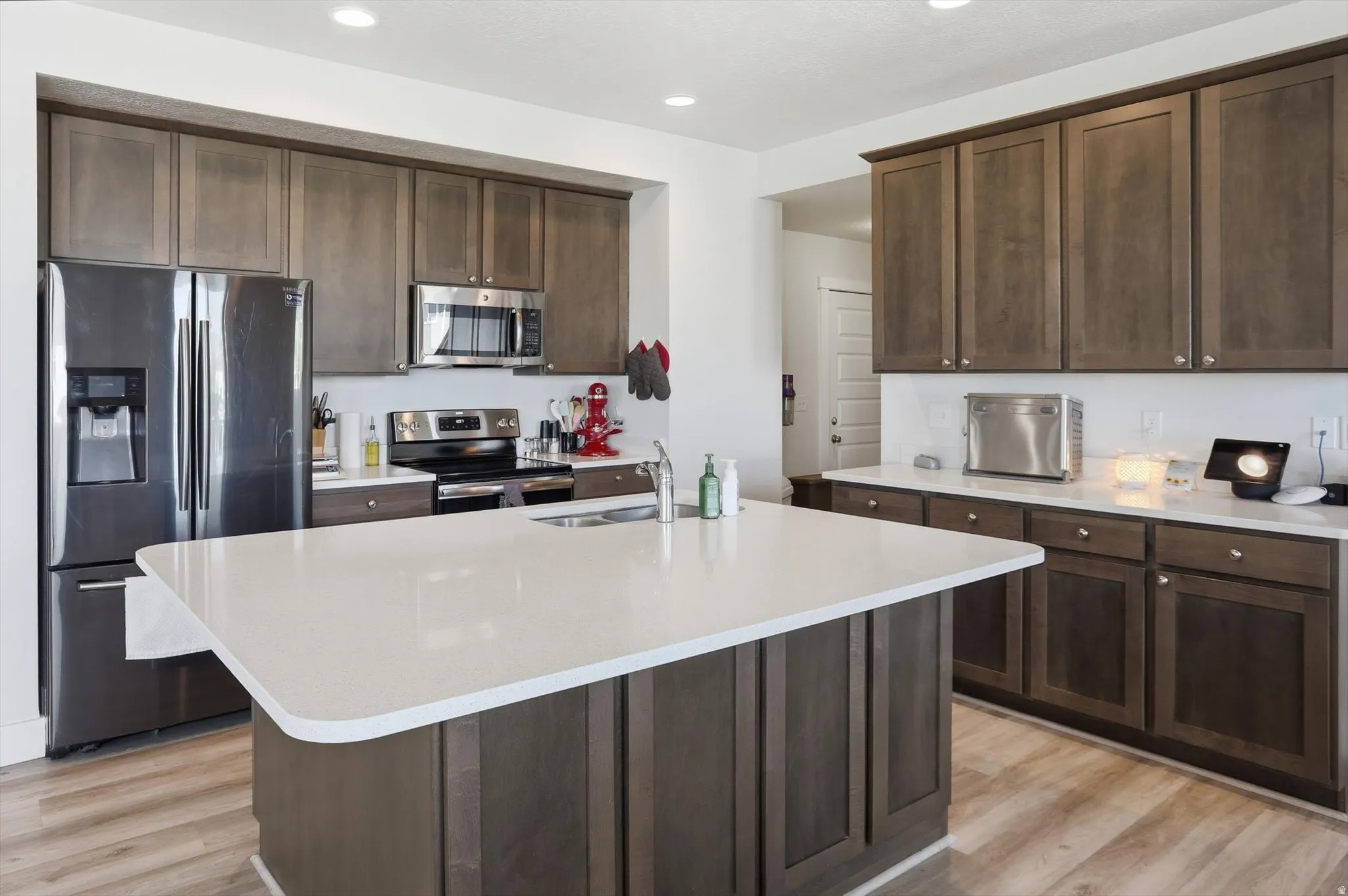Spacious kitchen featuring a large central island with a white countertop, dark wood-finish cabinetry, stainless steel appliances, and wood-finish flooring