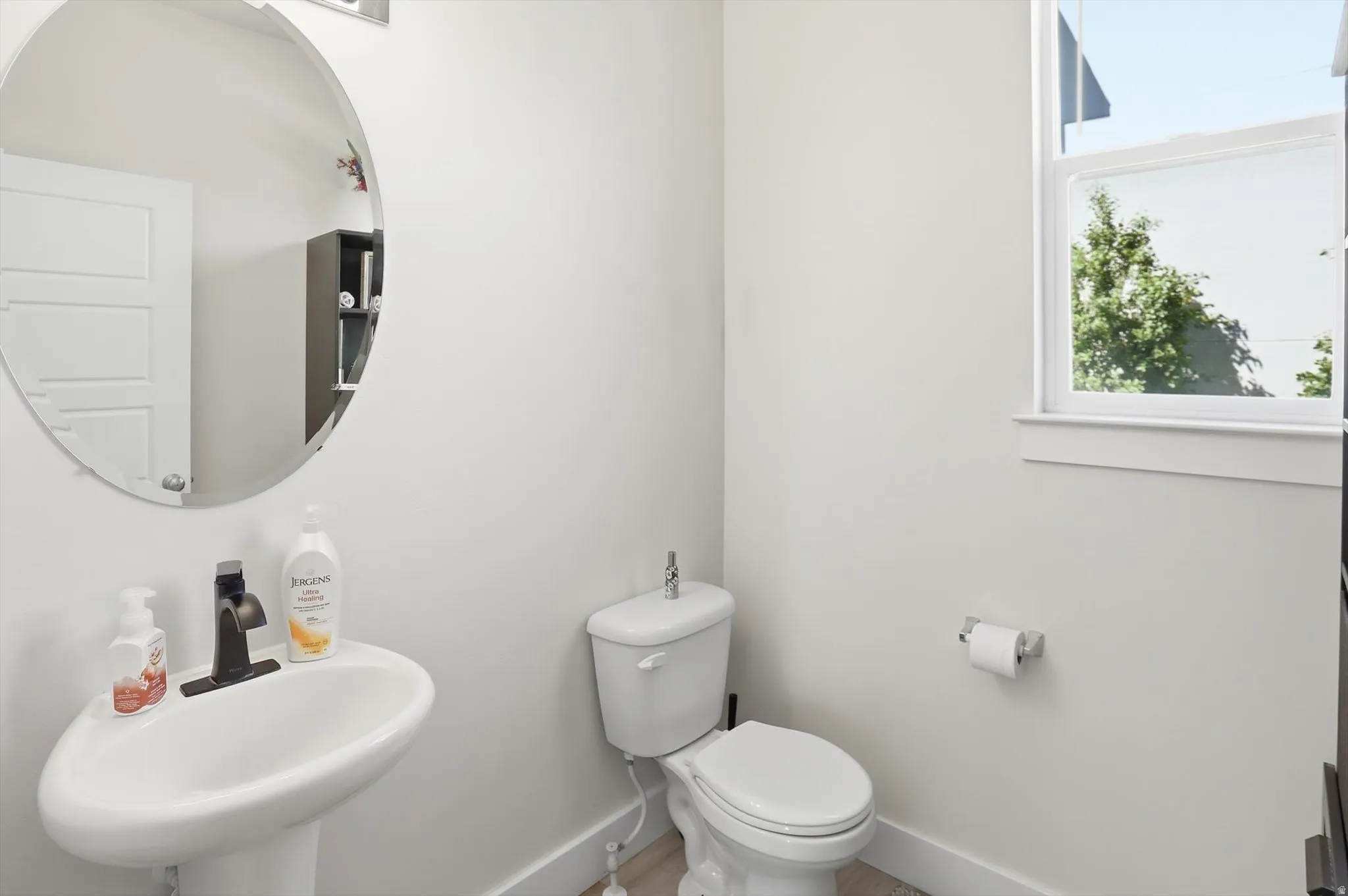 Powder room featuring a pedestal sink with a matte black faucet, a round wall-mounted mirror, a white toilet, and a window