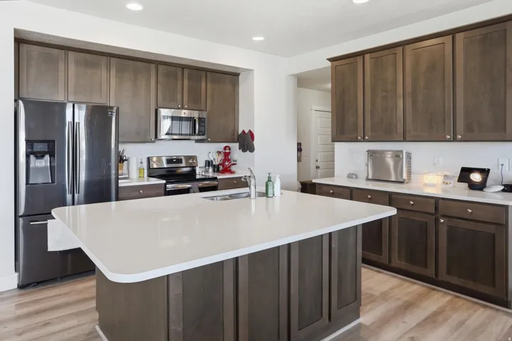 Spacious kitchen featuring a large central island with a white countertop, dark wood-finish cabinetry, stainless steel appliances, and wood-finish flooring