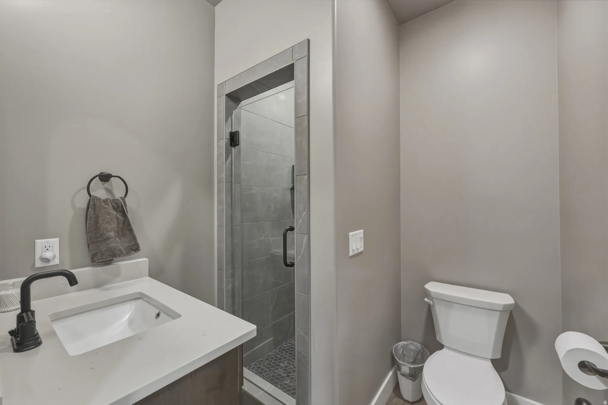 Modern bathroom featuring a glass-enclosed shower with gray tile surround and mosaic floor, a white vanity with integrated sink and dark bronze faucet, and light gray wall paint