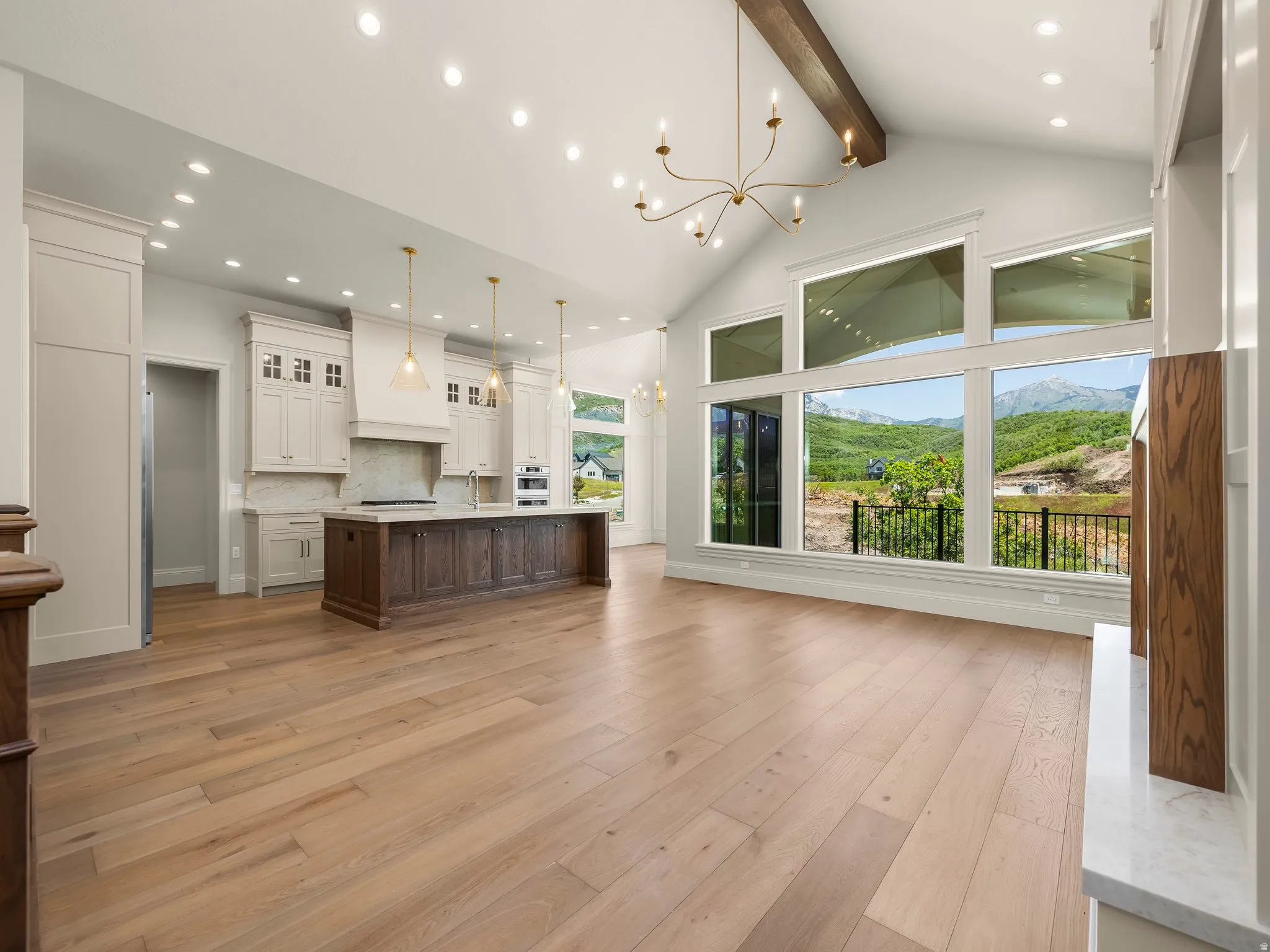 Open-concept living space featuring vaulted ceilings with an exposed wood beam, a large kitchen island with seating, extensive custom cabinetry, light wood-finish flooring, and a wall of windows showcasing mountain scenery