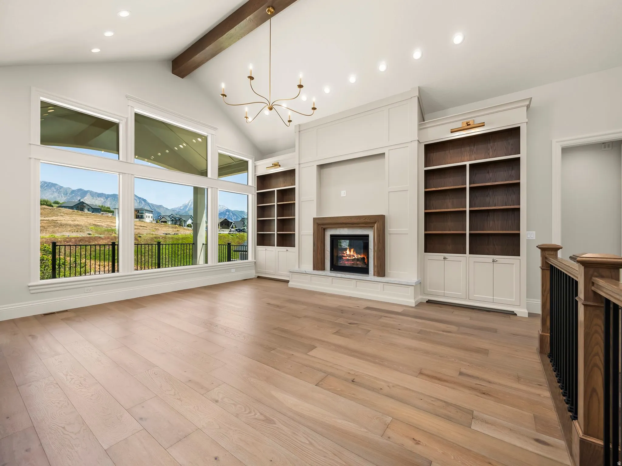 Spacious room featuring light wood-finish flooring, a vaulted ceiling with exposed beam and chandelier, a built-in fireplace with a wood and stone surround, custom built-in shelving, and large picture windows showcasing mountain scenery