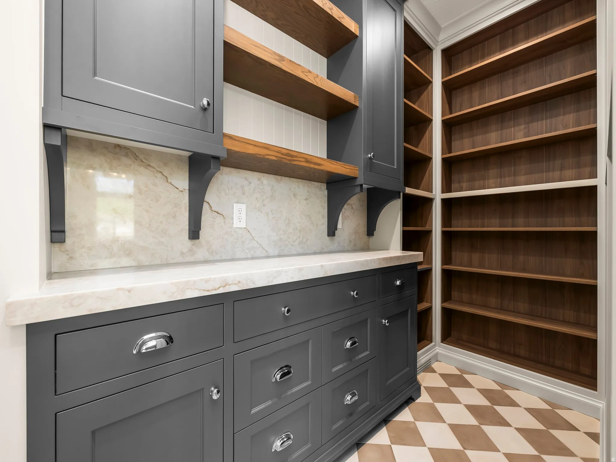Custom built-in pantry featuring extensive wood shelving, gray shaker-style cabinetry, light-toned stone countertops, a complementary stone backsplash, and a checkered tile floor