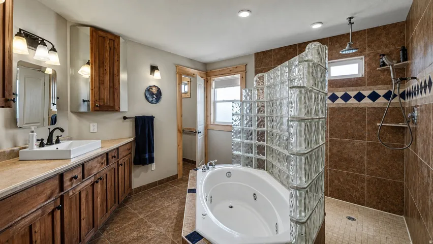 Spacious bathroom featuring a corner jetted tub, glass block shower enclosure, tiled shower surround, dual-light vanity fixture, and a wood-finish vanity with countertop sink