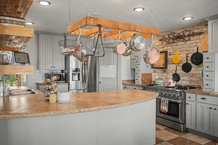 Spacious kitchen featuring a peninsula countertop, ample light-grey cabinetry, stainless steel appliances, a pot rack, and a brick-patterned accent wall