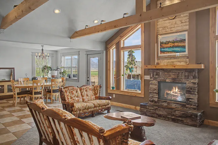 Living room featuring a stacked stone fireplace with wood mantel, large windows with wood trim, and wood beam ceiling accents