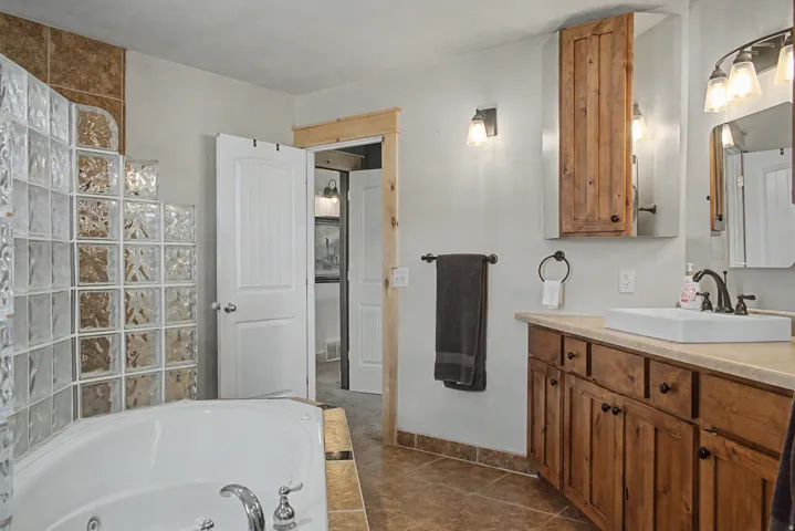 Bathroom featuring a corner soaking tub with a glass block surround, wood-finish vanity with an integrated sink, and tile flooring