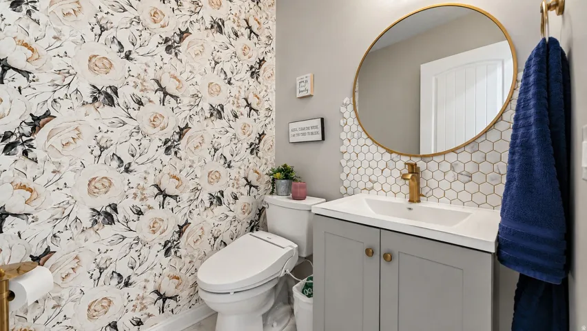 Powder room featuring a floral accent wall, a light gray vanity with gold-tone hardware, a white countertop, and a white hexagonal tile backsplash