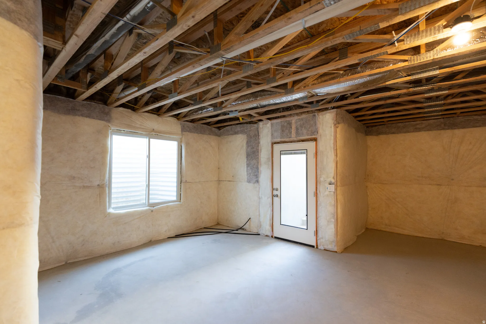 Unfinished basement featuring exposed wood ceiling joists, concrete flooring, insulated walls, a single-pane window with horizontal blinds, and a white door with a frosted glass insert