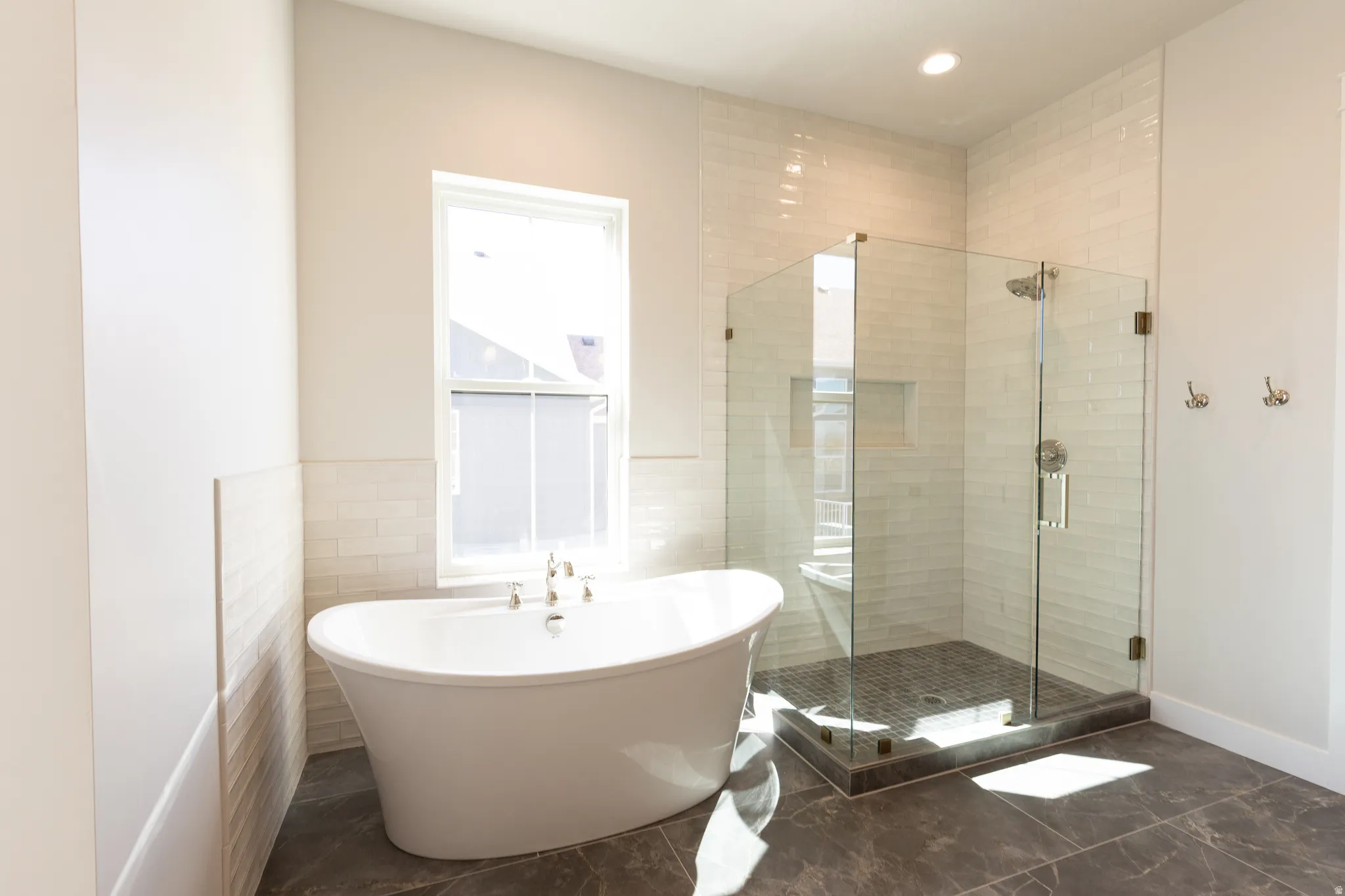 Spacious bathroom featuring a freestanding soaking tub, a clear glass enclosed shower, dark tile flooring, and white subway tile wainscoting