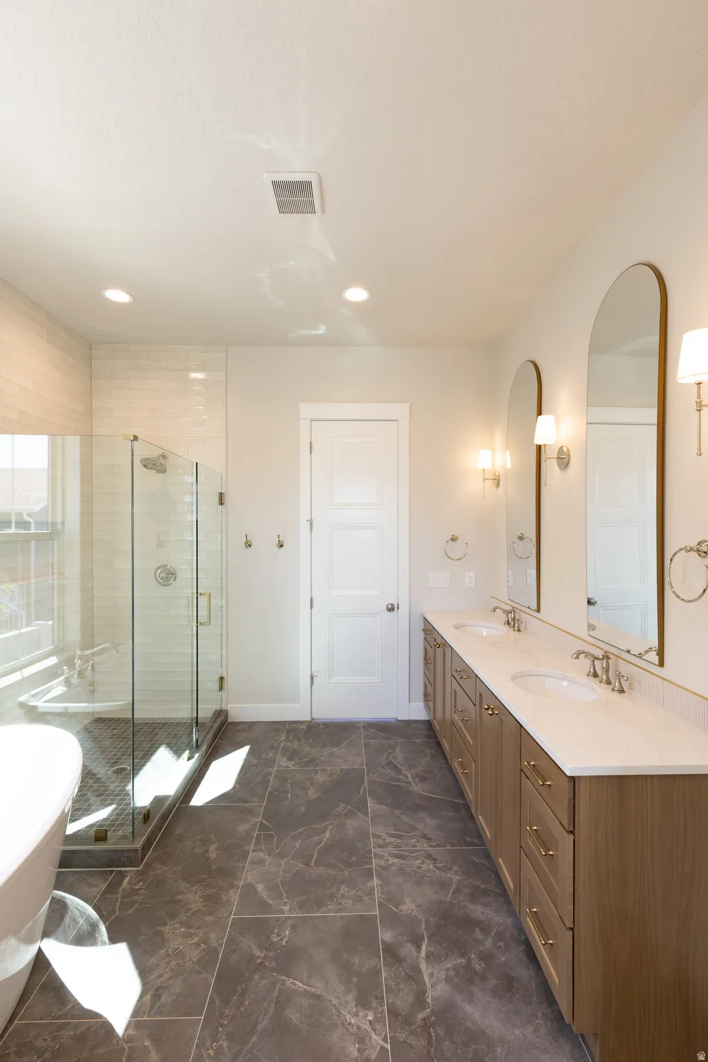 Spacious bathroom featuring a dual vanity with light wood-finish cabinetry, stone-look tile flooring, and a glass-enclosed shower with subway tile surround