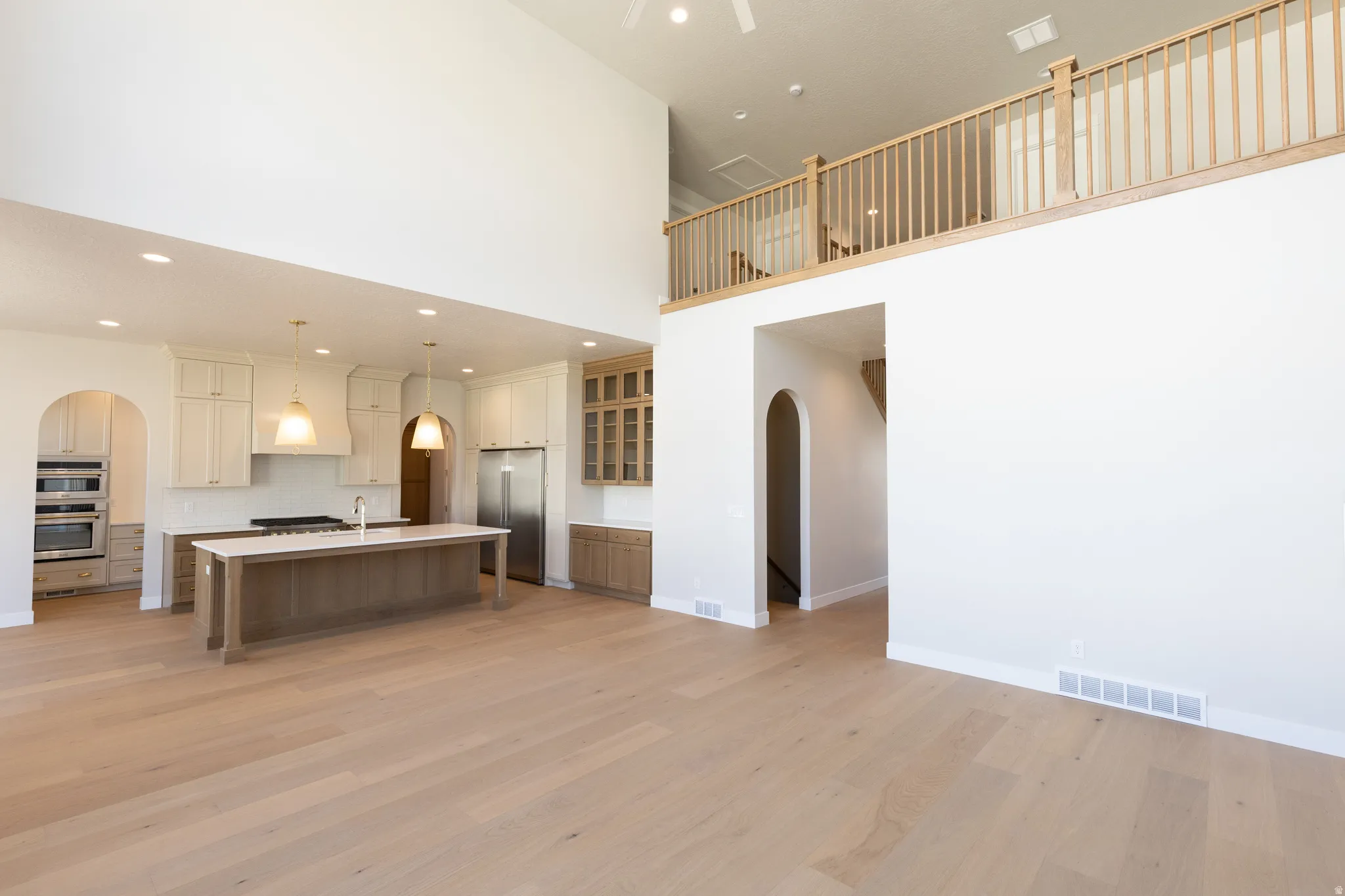 Spacious open-concept living area featuring light wood-finish flooring, a two-story ceiling, a gourmet kitchen with a central island, and an upper-level wood railing