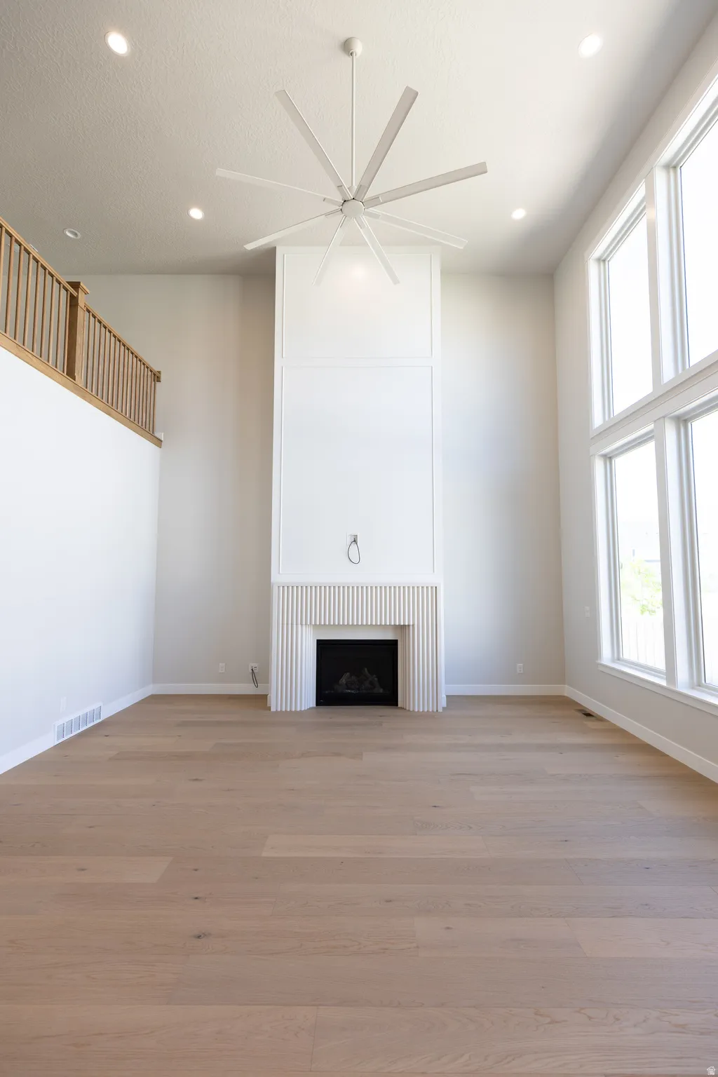Spacious living area featuring a floor-to-ceiling fireplace with reeded paneling, high ceilings with recessed lighting, a multi-blade ceiling fan, expansive windows, and light wood-finish flooring
