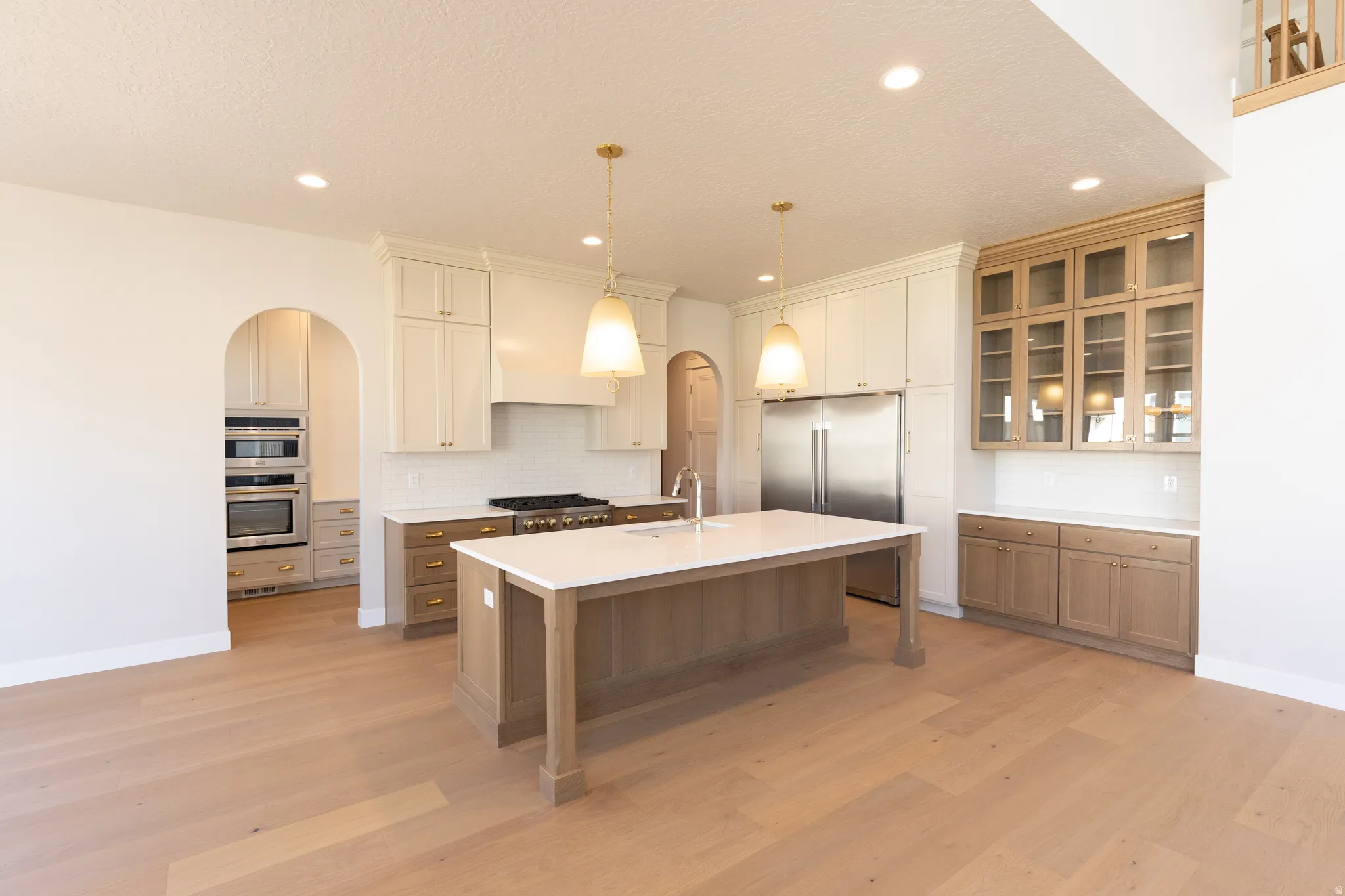 Gourmet kitchen featuring a large central island with a sink, stainless steel appliances, a gas range with an overhead vent hood, ample cabinetry in varied finishes, and light wood-finish flooring