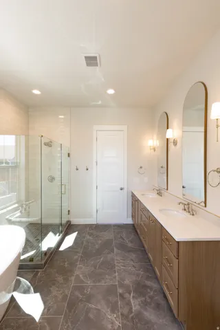 Spacious bathroom featuring a dual vanity with light wood-finish cabinetry, stone-look tile flooring, and a glass-enclosed shower with subway tile surround