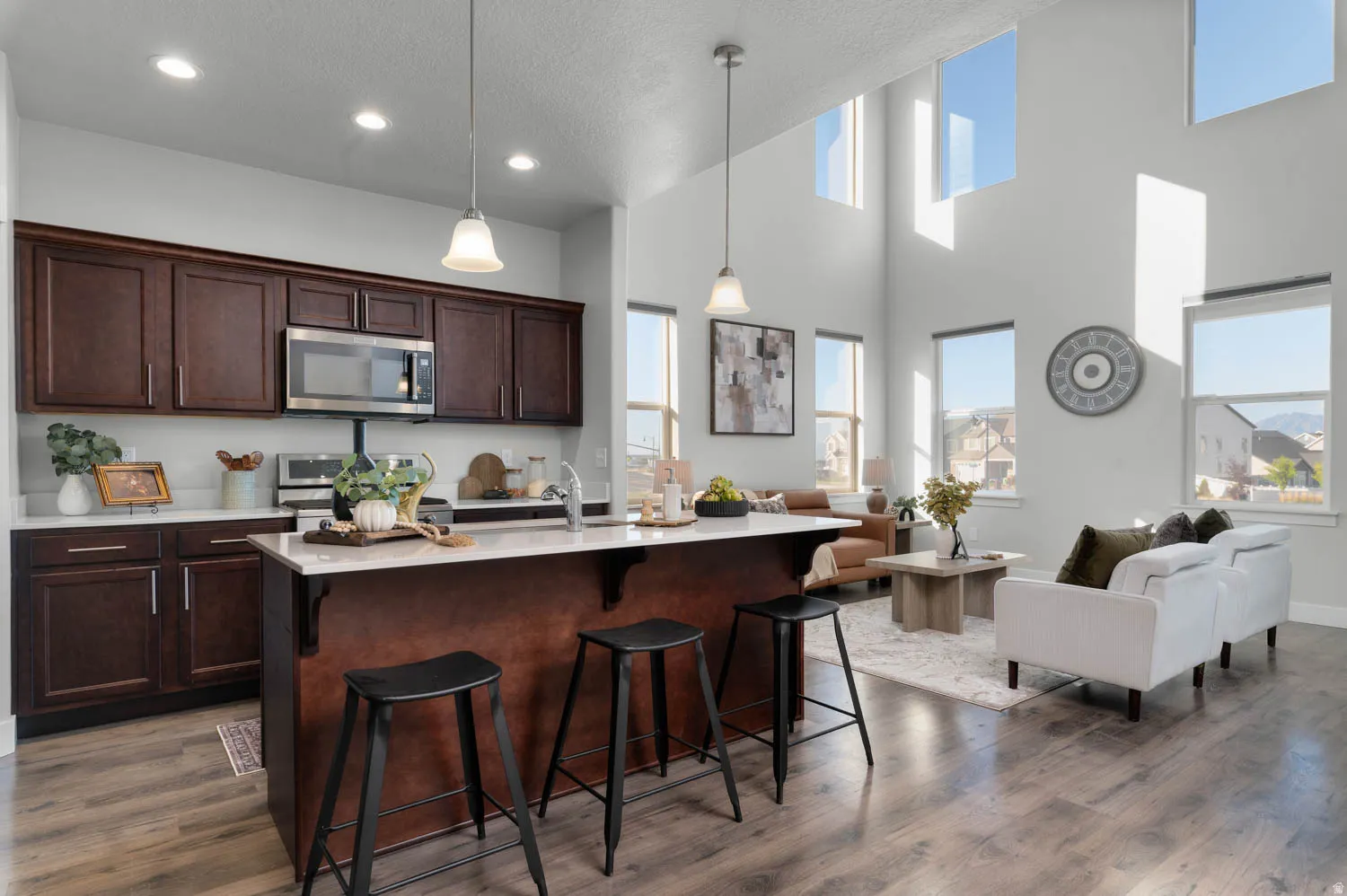 Open-concept living space featuring a spacious kitchen island with seating, dark wood-finish cabinetry, stainless steel appliances, and high ceilings with multiple windows