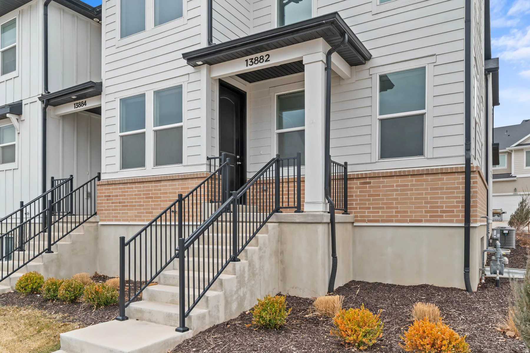 Multi-level townhouse exterior featuring light-toned horizontal siding, a covered entry with a black door, concrete steps with black metal railings, and a brick facade accent