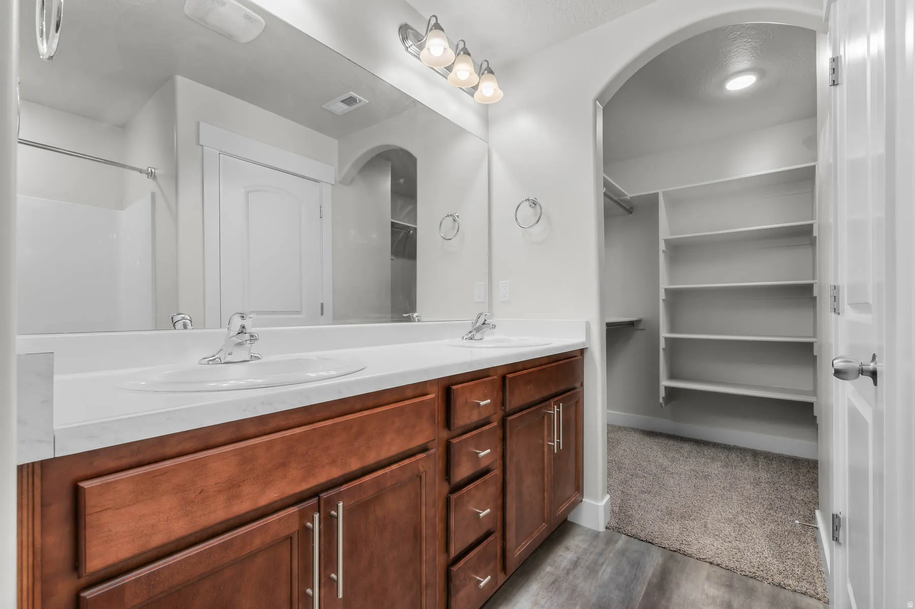 Bathroom vanity featuring dual sinks, a large mirror, wood-finish cabinetry with silver-tone hardware, and a walk-in closet with built-in shelving