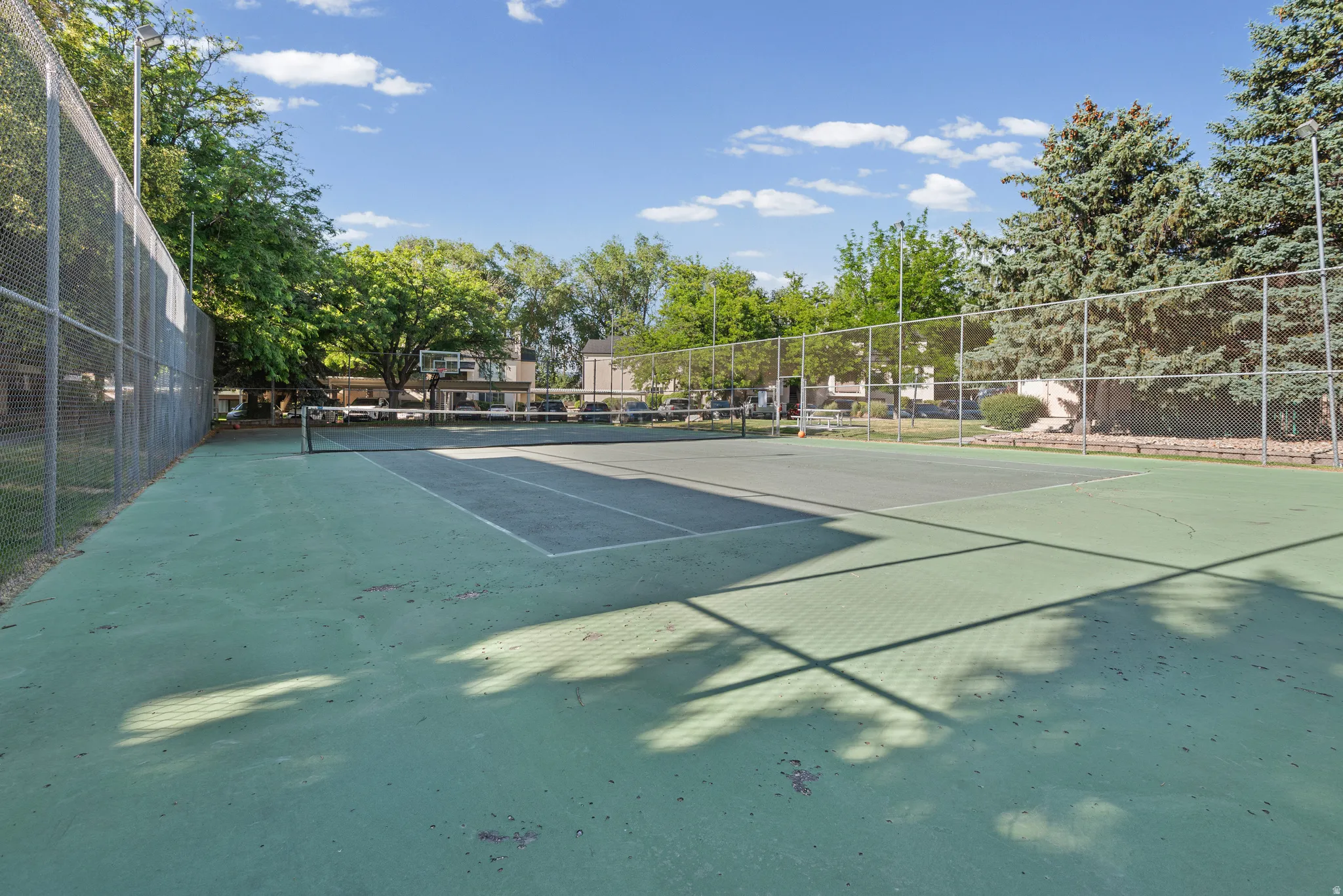 Community tennis court featuring a hard surface, perimeter chain-link fencing, and lush surrounding greenery