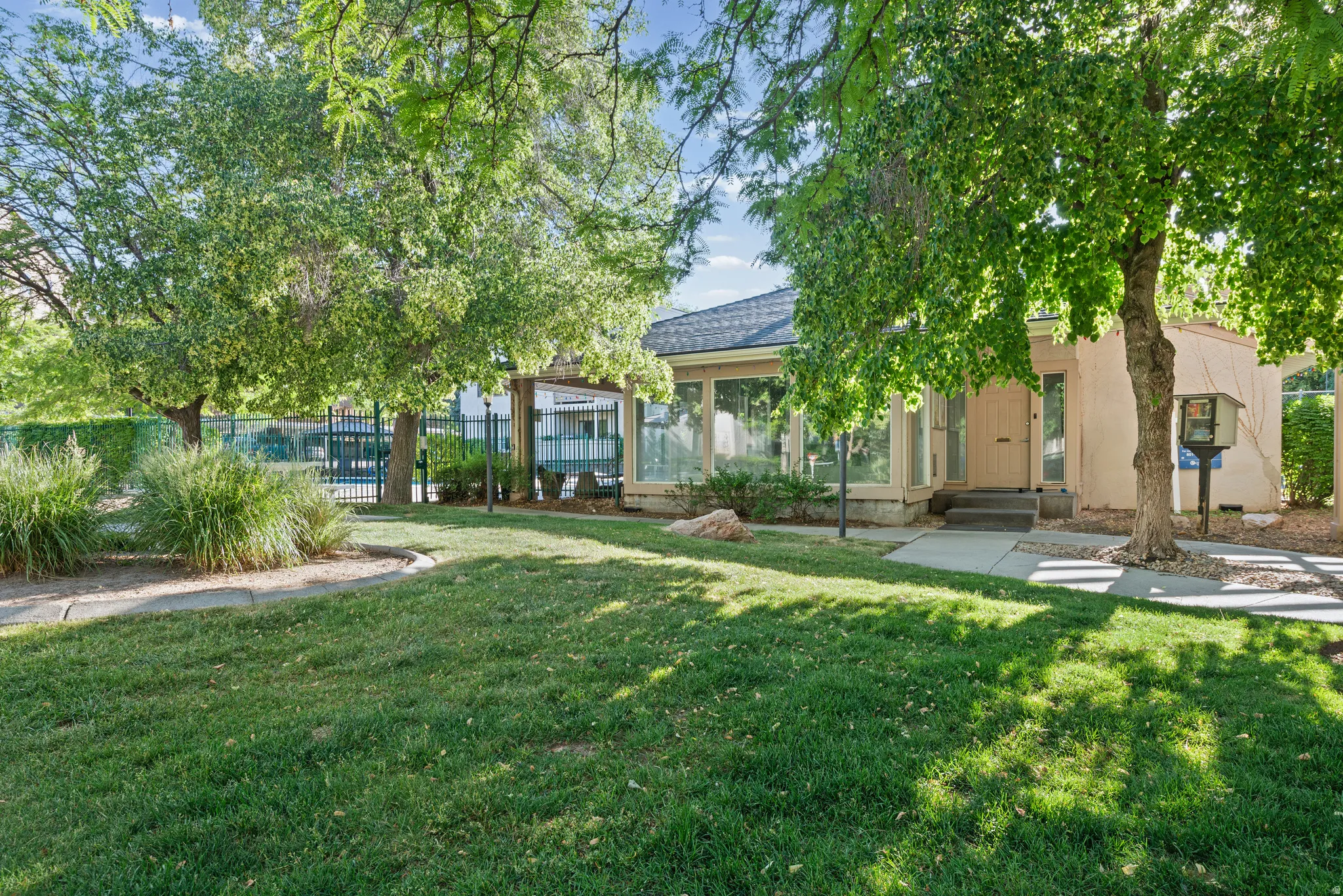 Lush green lawn with mature trees, a concrete walkway, and a beige exterior structure featuring large windows and a covered entrance