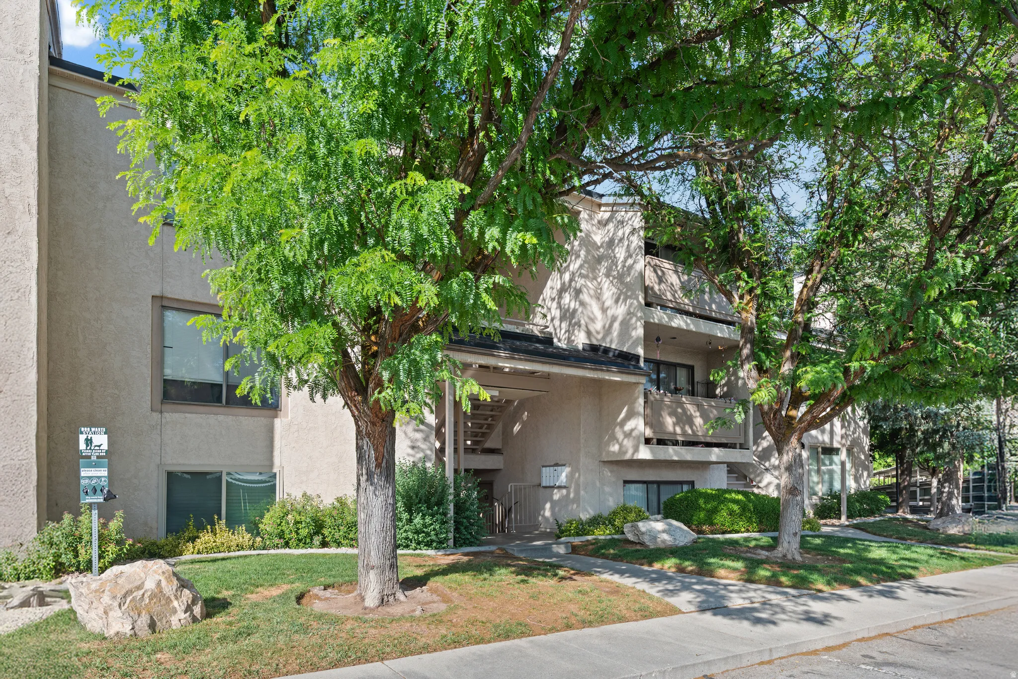 Multistory building with light-colored stucco exterior, multiple balconies, and mature trees providing natural shading