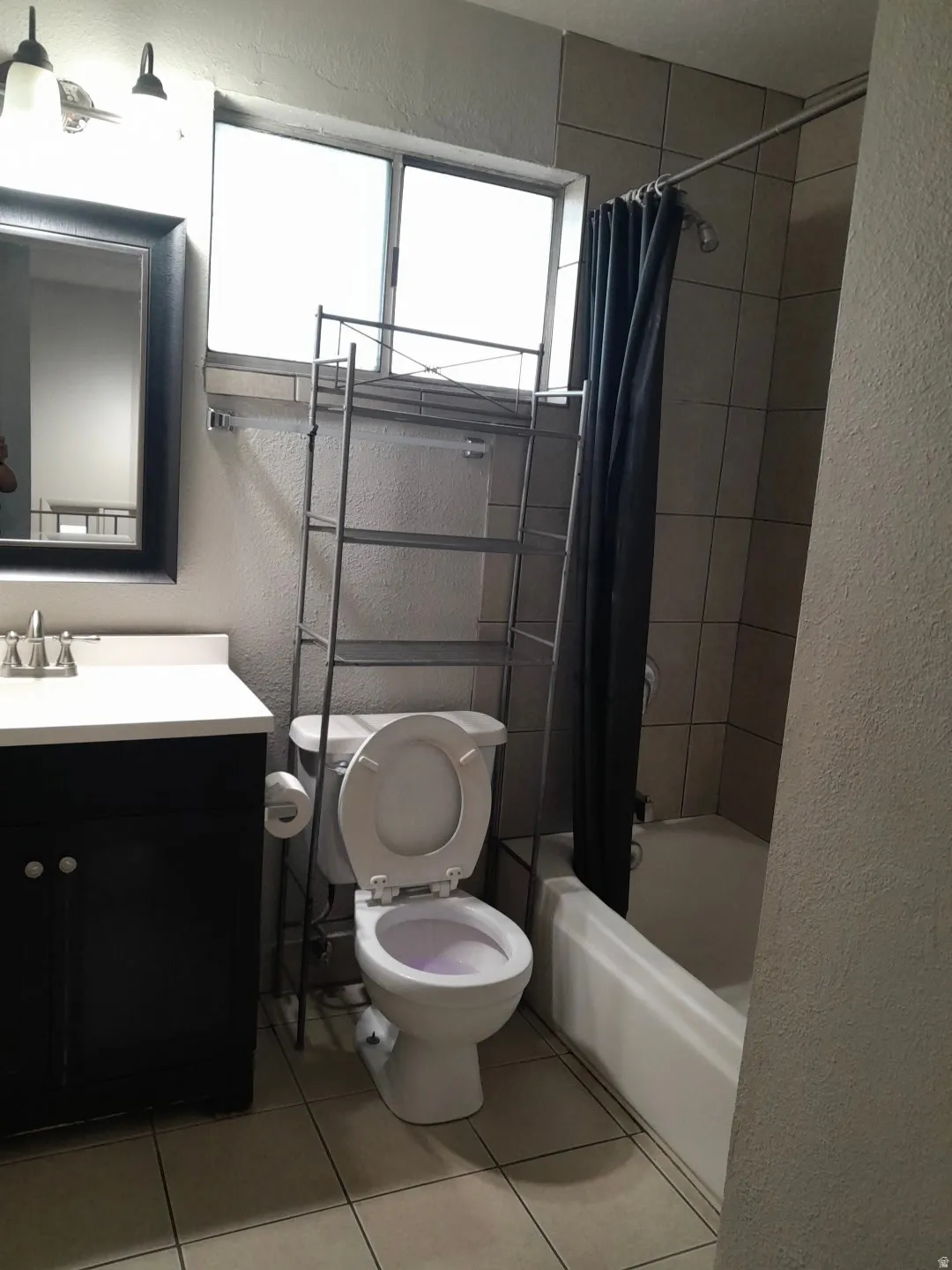 Bathroom featuring a white vanity with dark cabinetry, a framed mirror, an overhead light fixture, a window with obscure glass, a toilet, a tiled shower surround, and a white bathtub