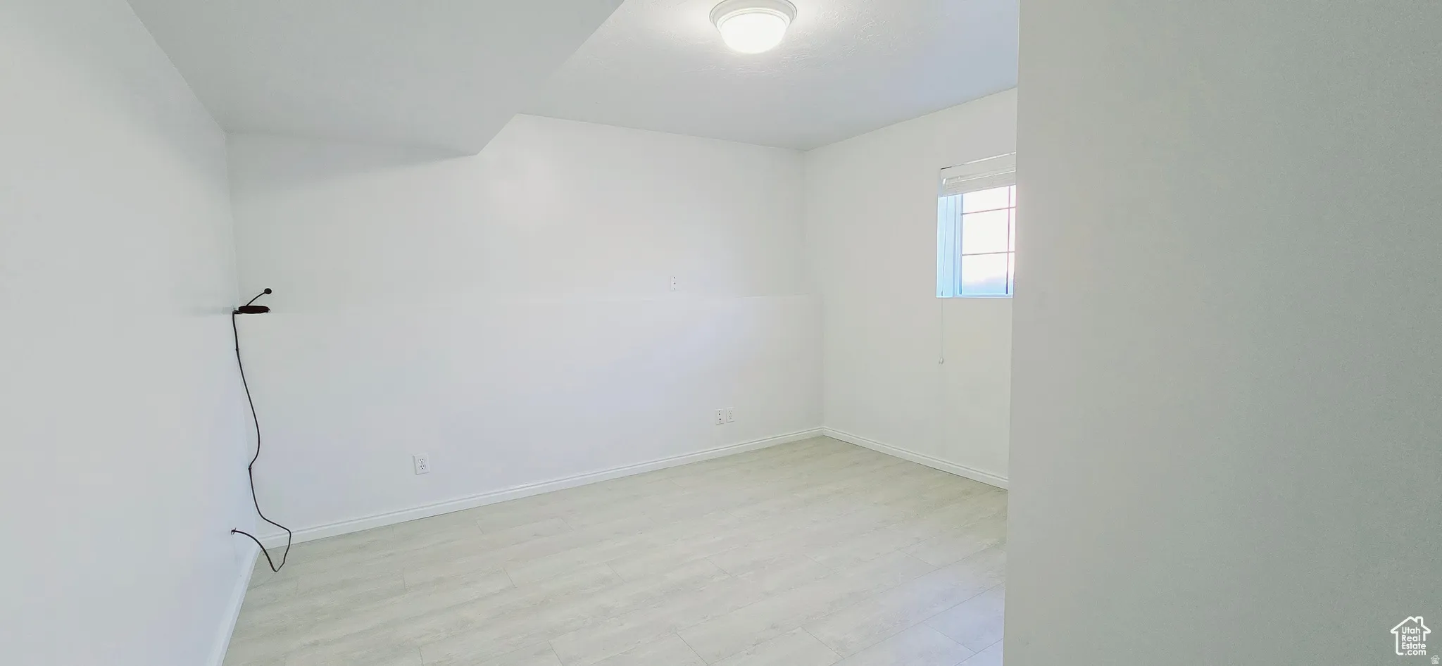 Light-filled room featuring neutral white walls, wood-finish flooring, a single window with blinds, and a flush-mount ceiling light fixture