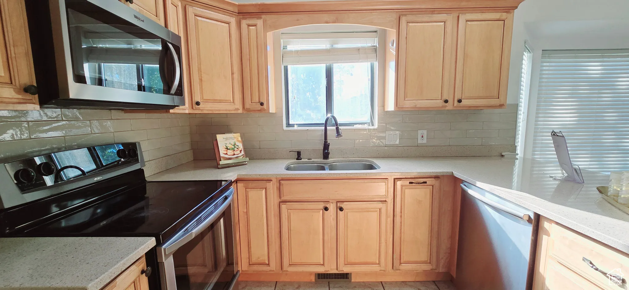 Kitchen featuring light wood cabinetry, light countertops, subway tile backsplash, stainless steel appliances, and a double basin sink with a dark-finish faucet