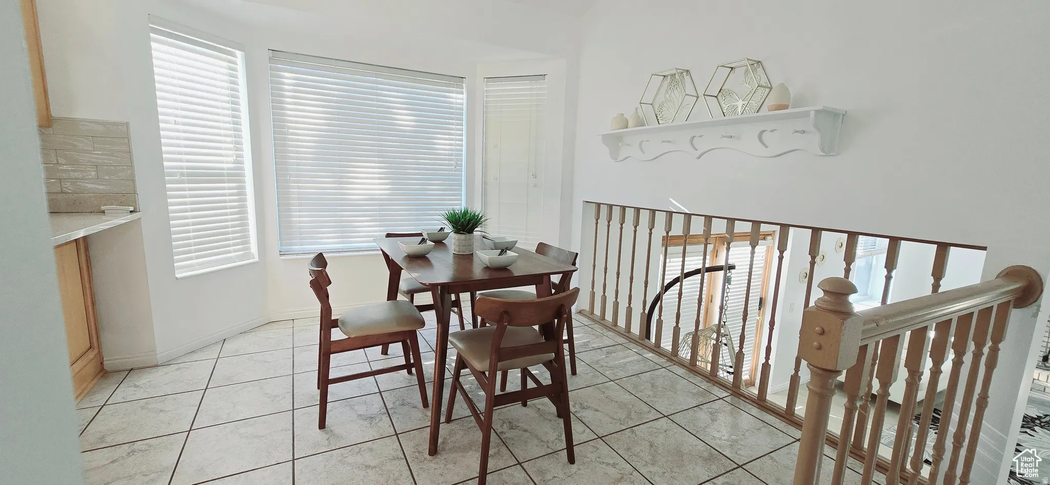 Bay window with horizontal blinds, neutral tone ceramic tile flooring, built-in counter with light wood cabinetry, white wall shelving, and wooden stair railing