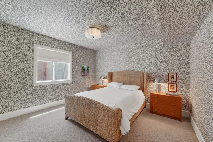 Interior room featuring patterned wallpaper, a white-framed window with a roller shade, and light-colored carpeting