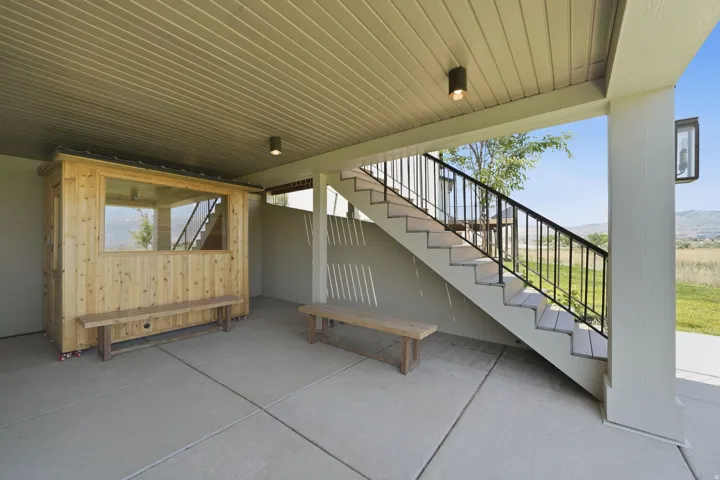 Covered concrete patio featuring a built-in wooden structure with large windows, an exterior staircase with black metal railings, and dual exterior wall sconces