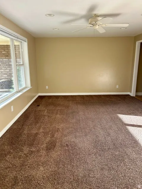 Spacious carpeted room featuring a ceiling fan, recessed lighting, neutral wall tones, white baseboards, and a large window with white trim