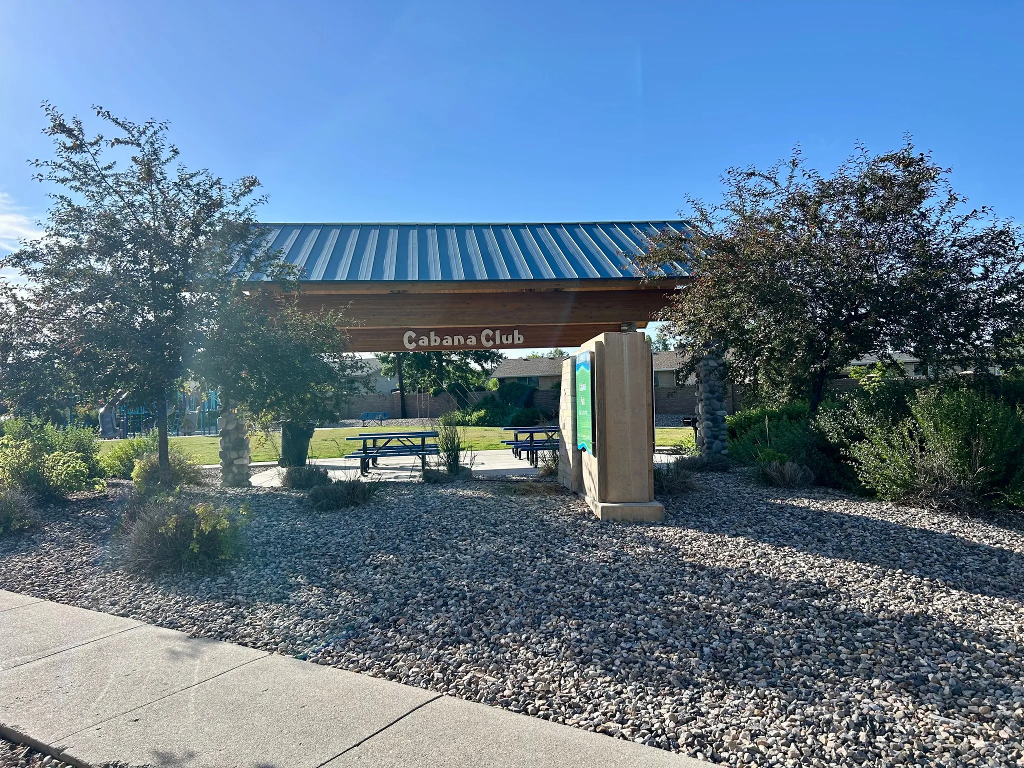 "Cabana Club" entrance featuring a corrugated metal roof over a wood beam structure, stone column accents, and surrounding landscaping with mature trees and ground cover
