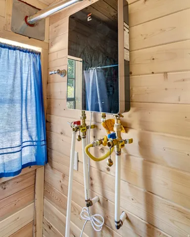 Tankless water heater with exposed brass and white PVC plumbing against natural wood-finish wall paneling