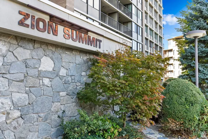 Exterior stone wall with raised signage, framed by mature green landscaping and a tall, multi-story building featuring individual balconies