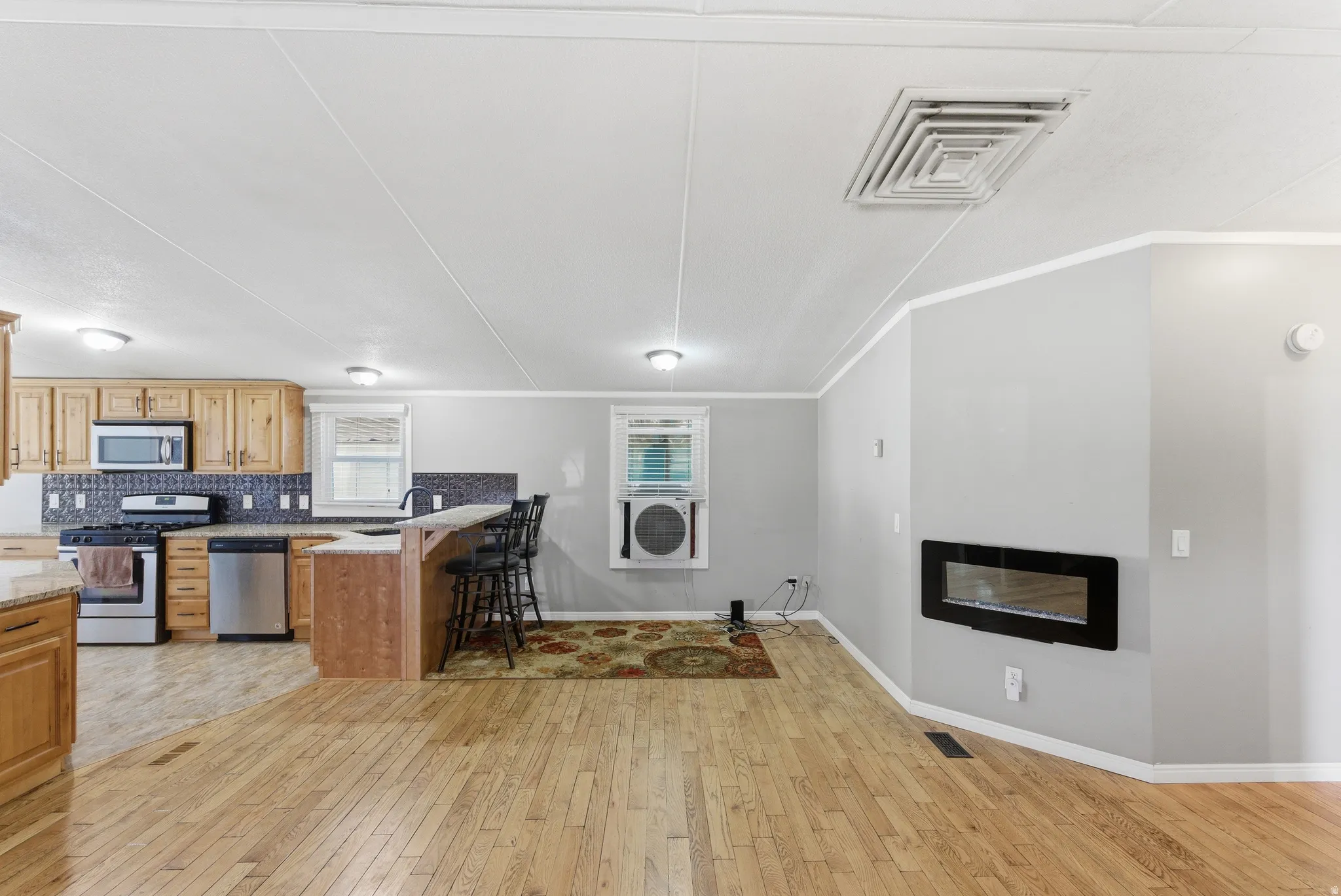 Open-concept living space featuring wood-finish flooring, a contemporary wall-mounted fireplace, a kitchen with wood cabinetry, stainless steel appliances, and a breakfast bar with stone-finish countertops