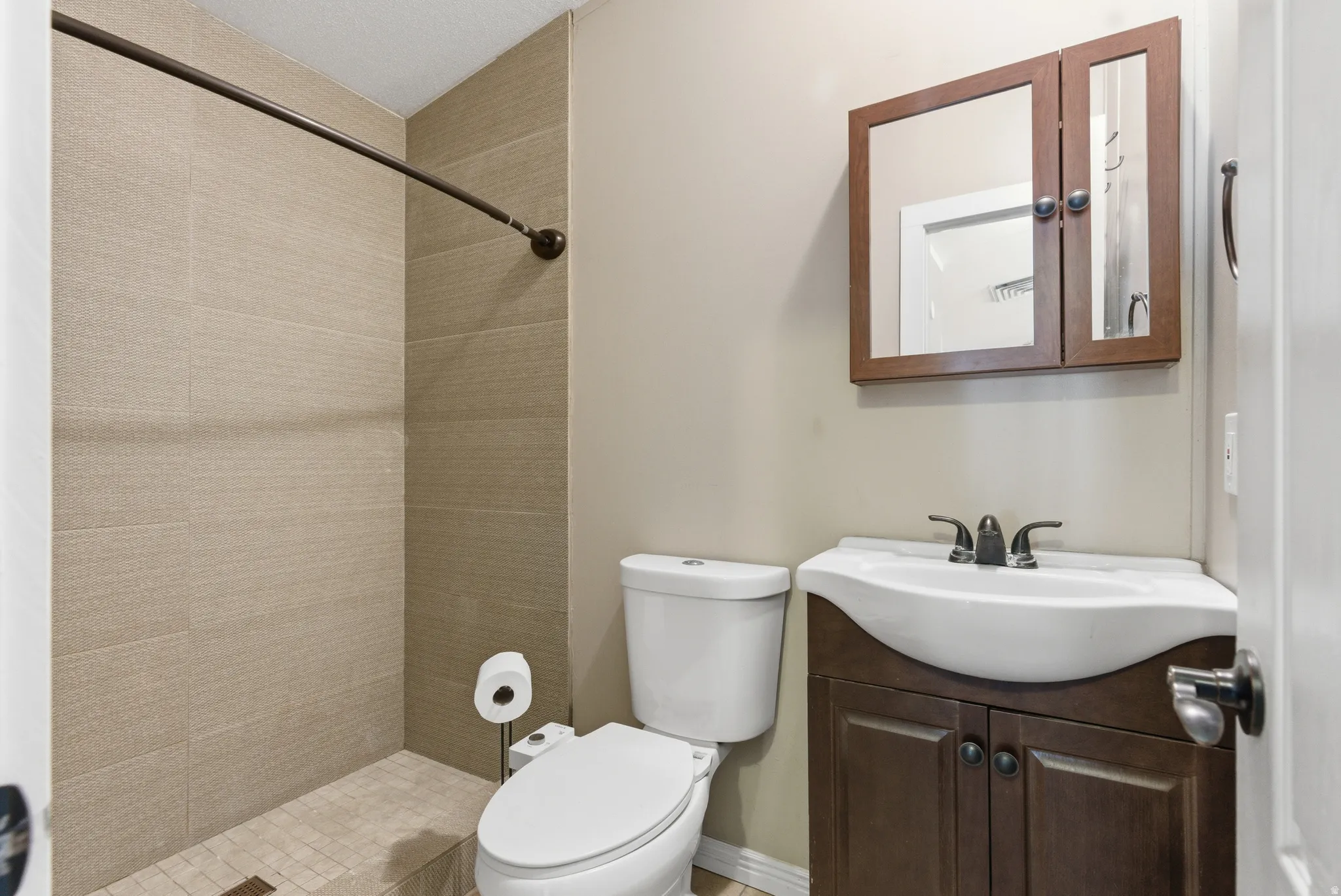 Bathroom featuring a walk-in shower with textured tile surround and mosaic tile flooring, a dark wood-finish vanity with an integrated white sink, a dark wood-finish mirrored medicine cabinet, and a toilet