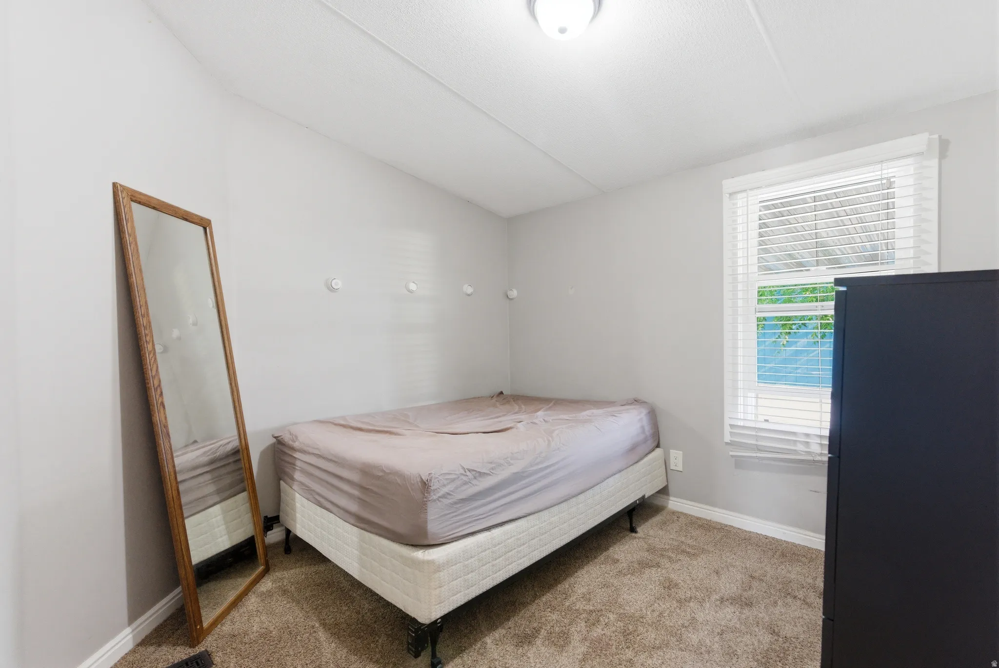 Carpeted room with a ceiling-mounted light fixture, a window with horizontal blinds, and a built-in wall shelf