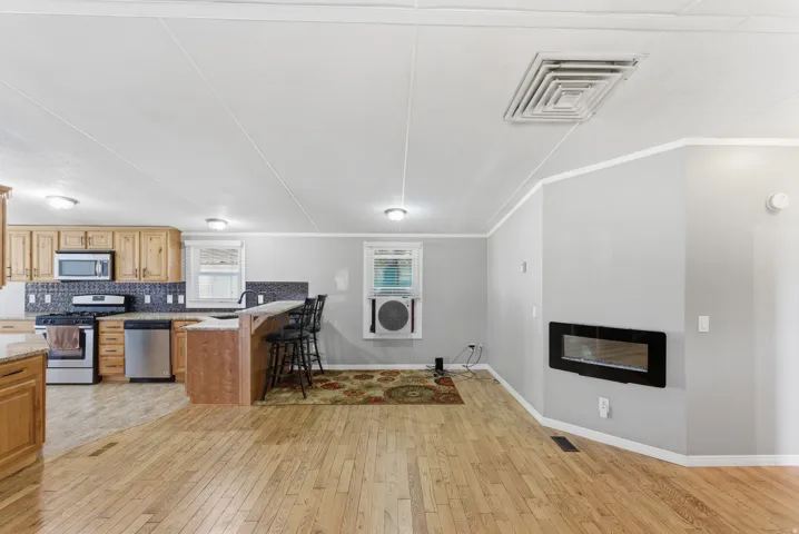 Open-concept living space featuring wood-finish flooring, a contemporary wall-mounted fireplace, a kitchen with wood cabinetry, stainless steel appliances, and a breakfast bar with stone-finish countertops