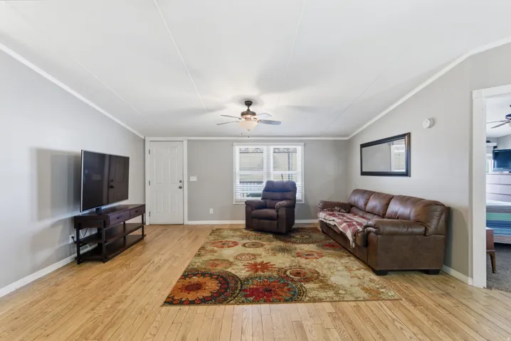 Living area featuring wood-finish flooring, light gray wall paint, a white paneled entry door, a ceiling fan with integrated lighting, and multiple windows