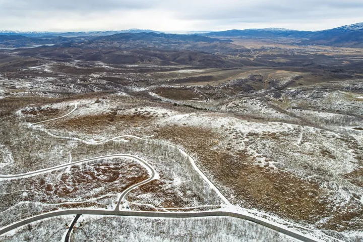 Expansive aerial landscape featuring a mountainous terrain with distant peaks, winding roads, and scattered tree cover