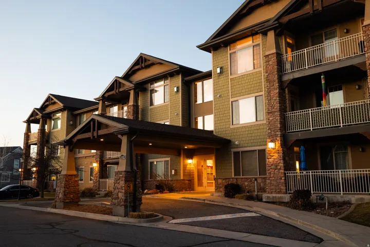 Building exterior featuring a covered entryway, stone facade accents, shingle siding, multiple balconies with metal railings, and integrated exterior lighting